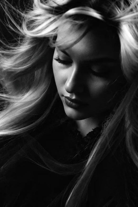 Black and white portrait of a woman with long flowing hair, wearing a dark sheer garment against a dark background.